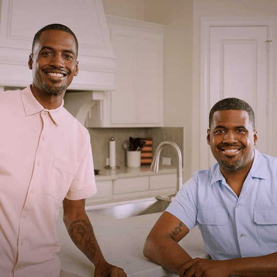 Two adult African-American men are standing in a white colored home kitchen.