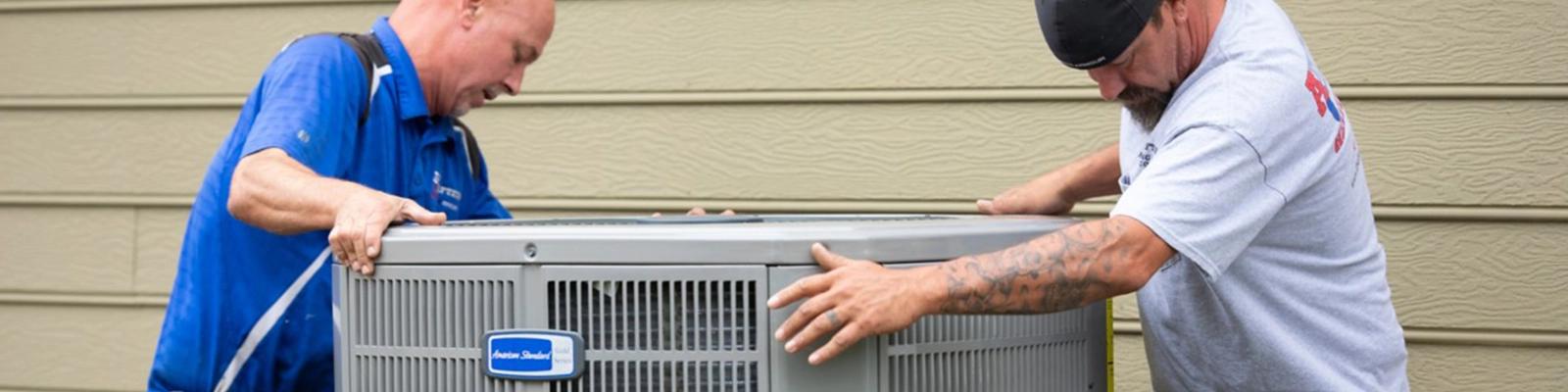 Two men are moving an American Standard Air outdoor unit.