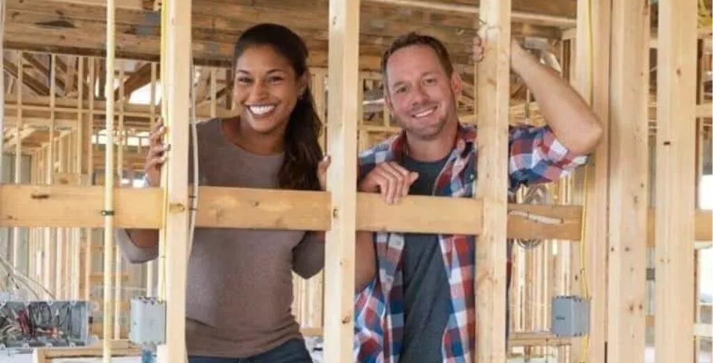 Brian and Mika Kleinschmidt stand smiling surrounded by wood beams inside of a home being renovated.