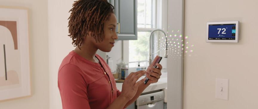 A woman stands in front of her American Standard smart thermostat and controls the temperature via her smart phone
