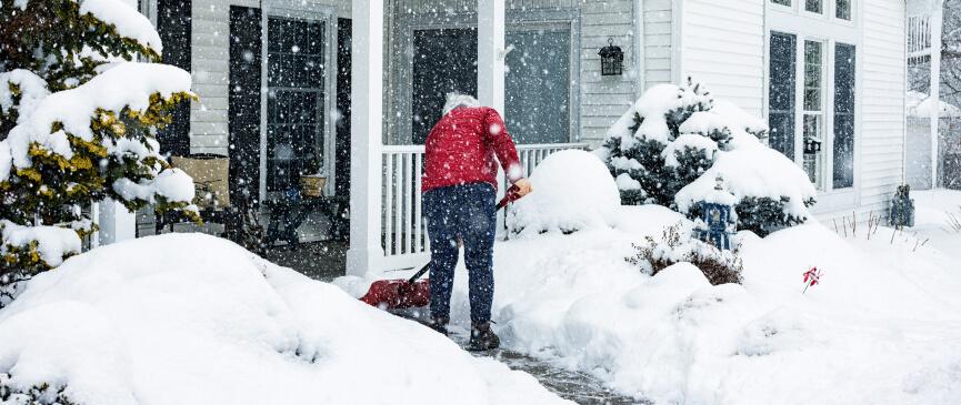 A person in a red coat shovels their walkway as snow falls in the winter