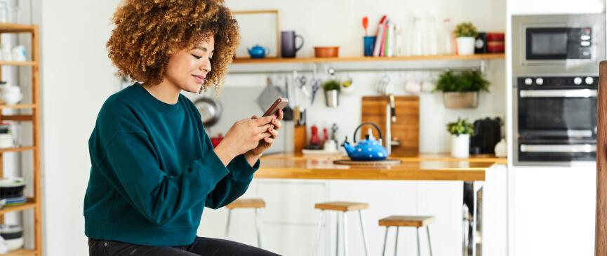 A woman sits in her kitchen, looking at her phone and preparing for a visit from her local HVAC dealer.