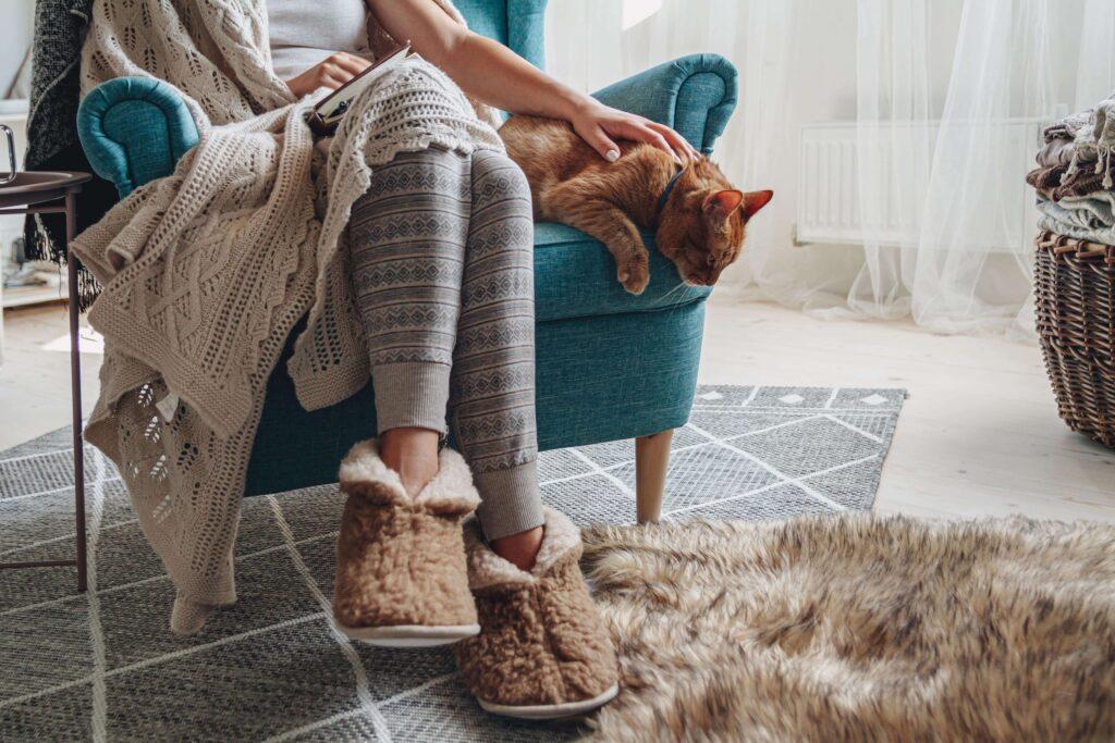 Woman sits with on a comfortable blue chair with a blanket while she pets a cat.