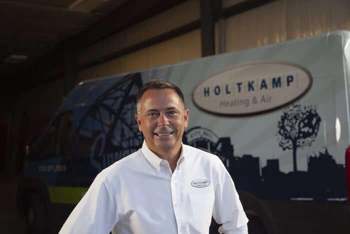 Matthew Holtkamp from Holtkamp Heating &amp; Air stands in front of a sign displaying the company logo.