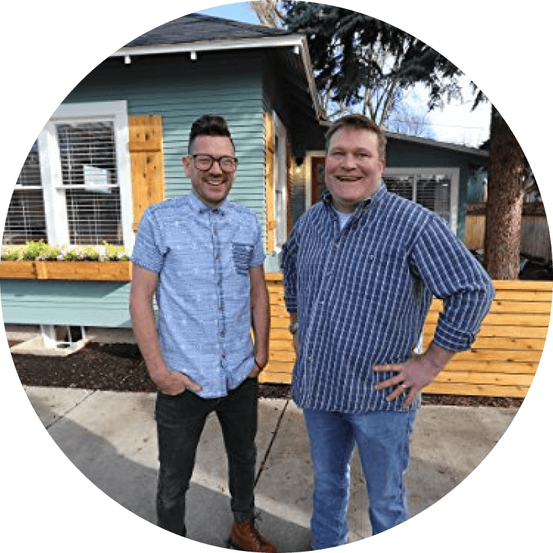 Two caucasian men are standing in front of a blue house.