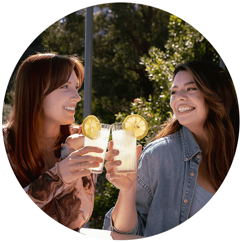 Two young caucasian women are sitting on a porch drinking lemonade.