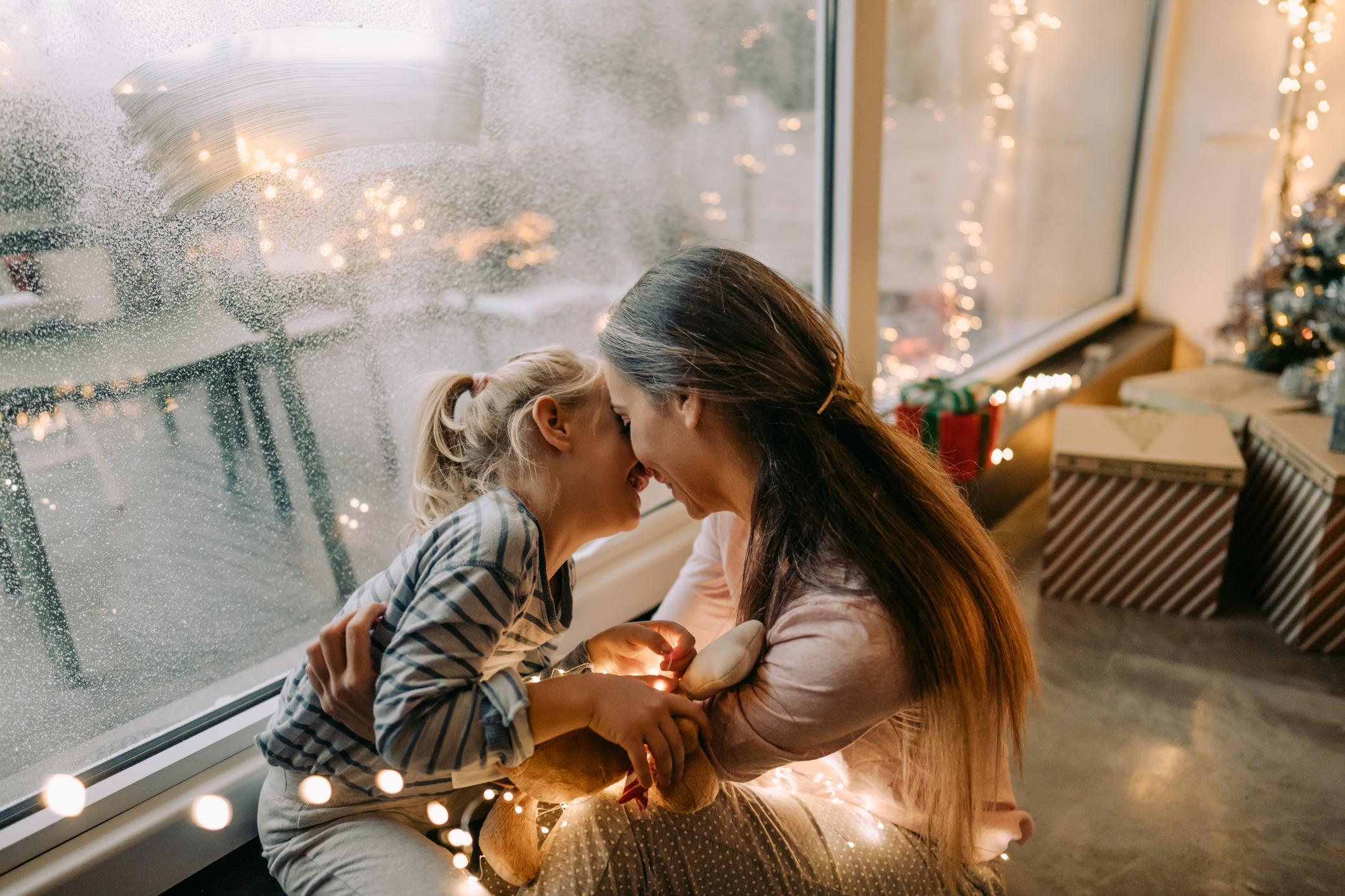 A young Caucasian woman is hugging her daughter by an indoor window with Christmas lights.